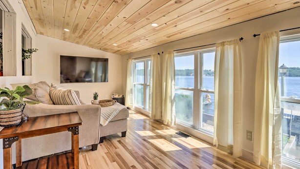 A cozy living room with a wooden ceiling, large windows with curtains, a sofa, a coffee table, and a TV mounted on the wall. - Dock & Boat Slip! Waterfront Lake Ozark Home (Lake Ozark)