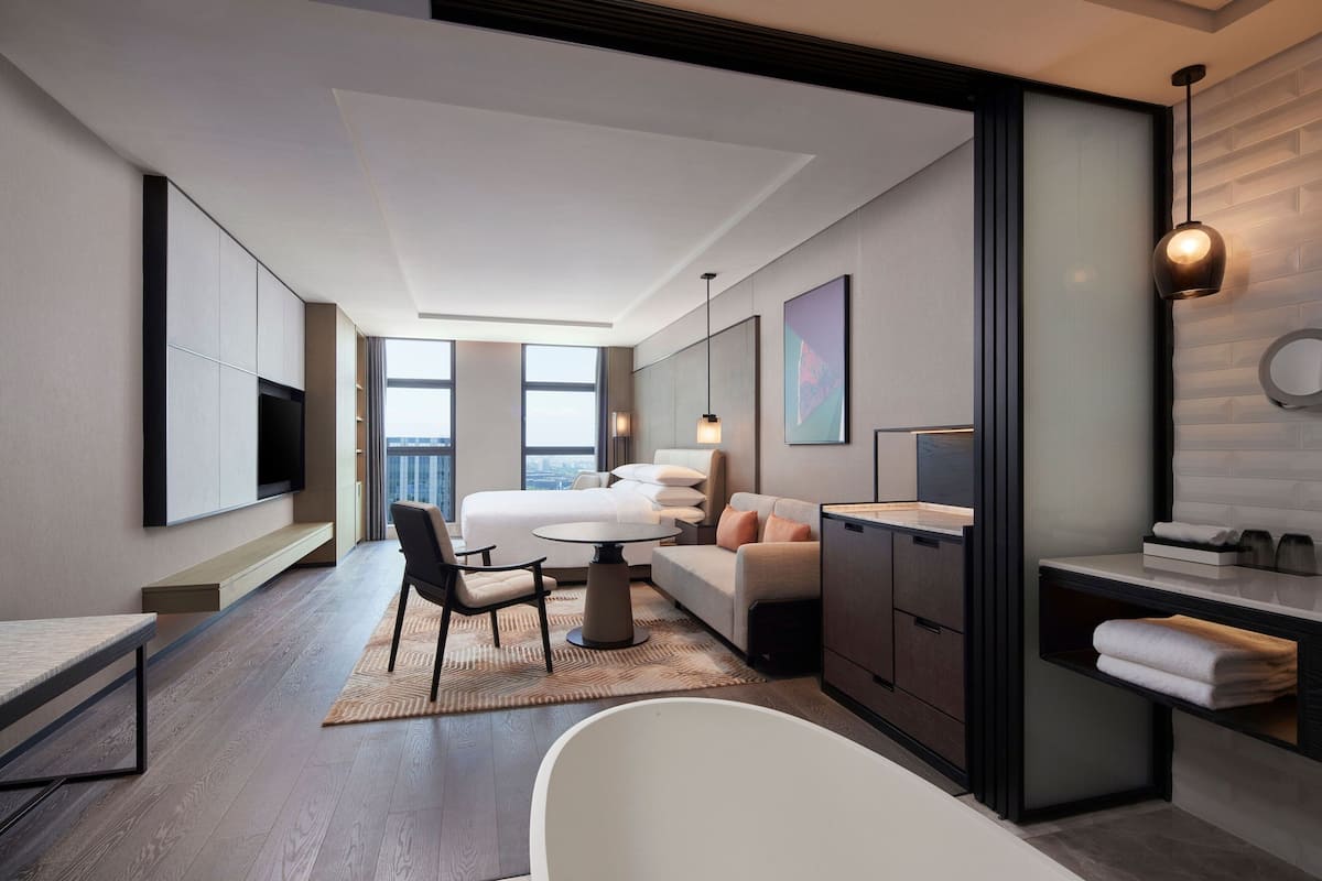 A modern hotel room with a large bed, a sitting area with a sofa and chairs, a dining table, a bathtub, and a TV mounted on the wall.