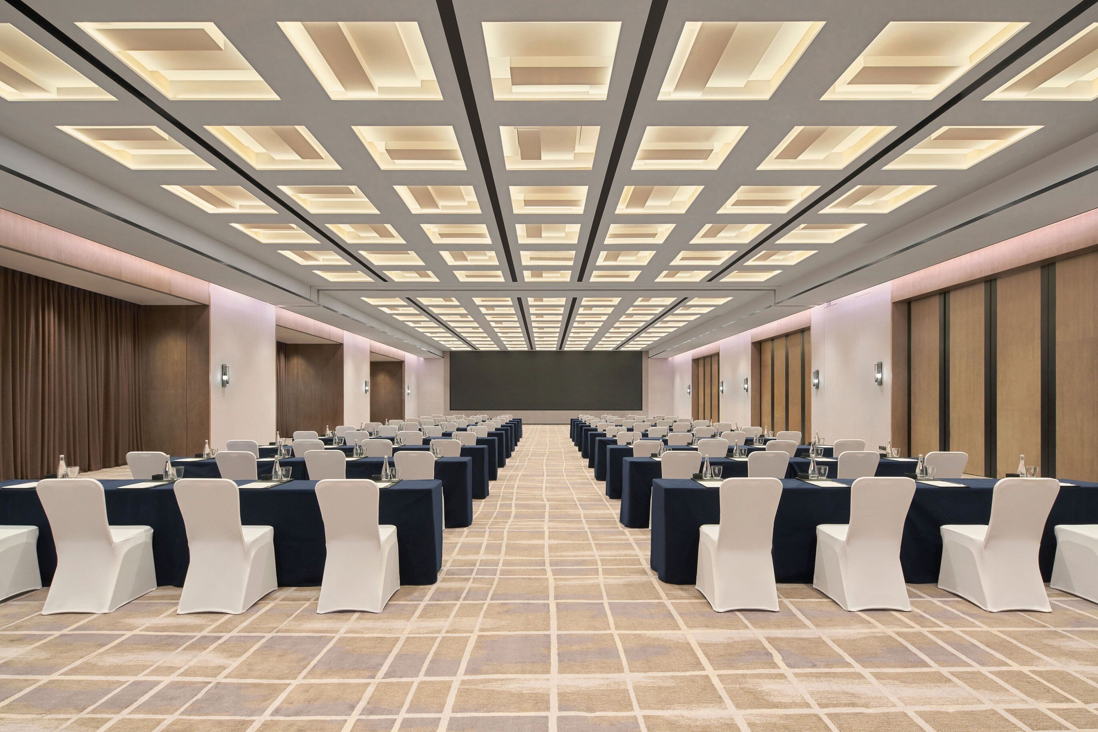 A conference room with rows of tables and chairs, a large screen at the front, and a patterned floor.