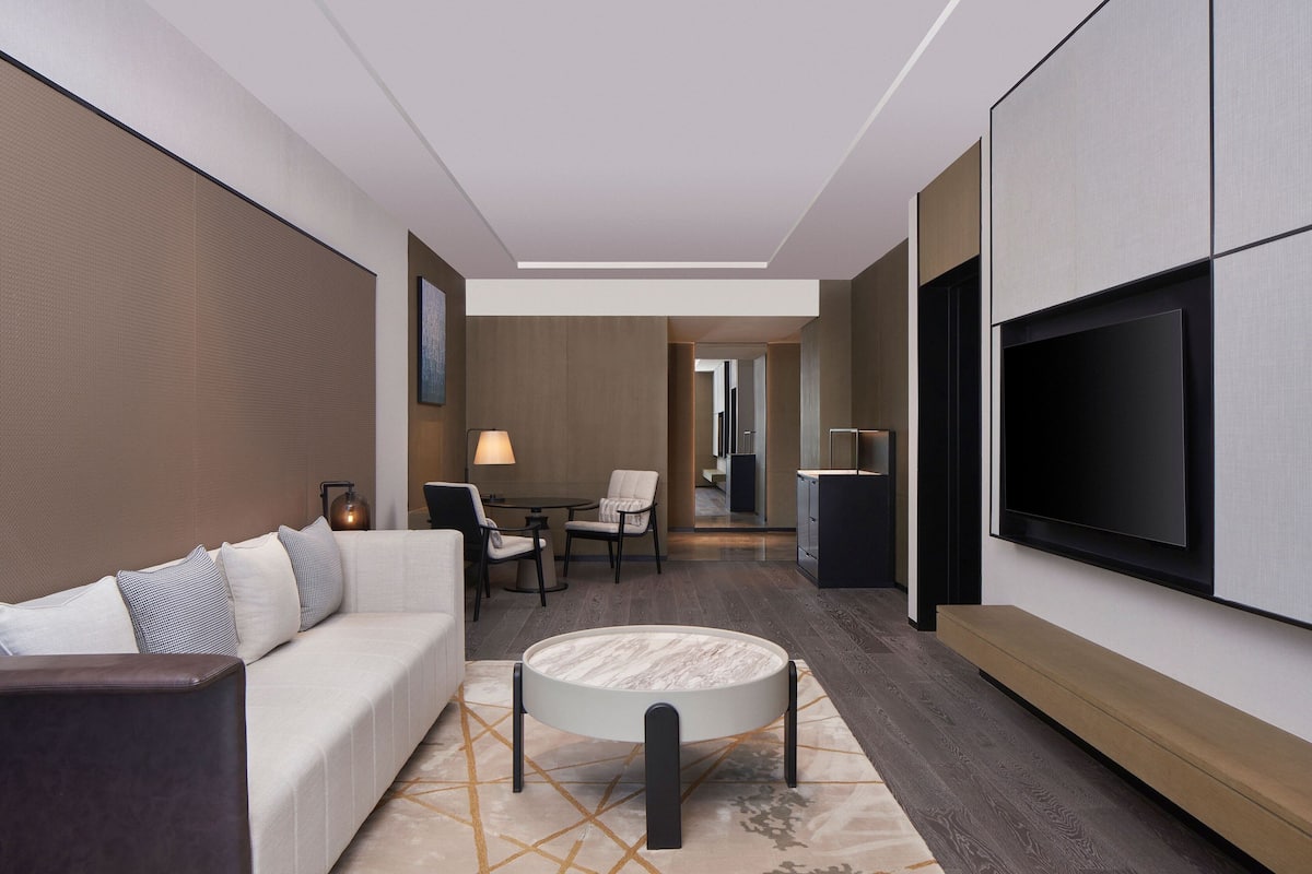 A modern hotel room with a sofa, a round coffee table, a flat-screen TV mounted on the wall, a dining area with a table and chairs, and a hallway leading to another room.