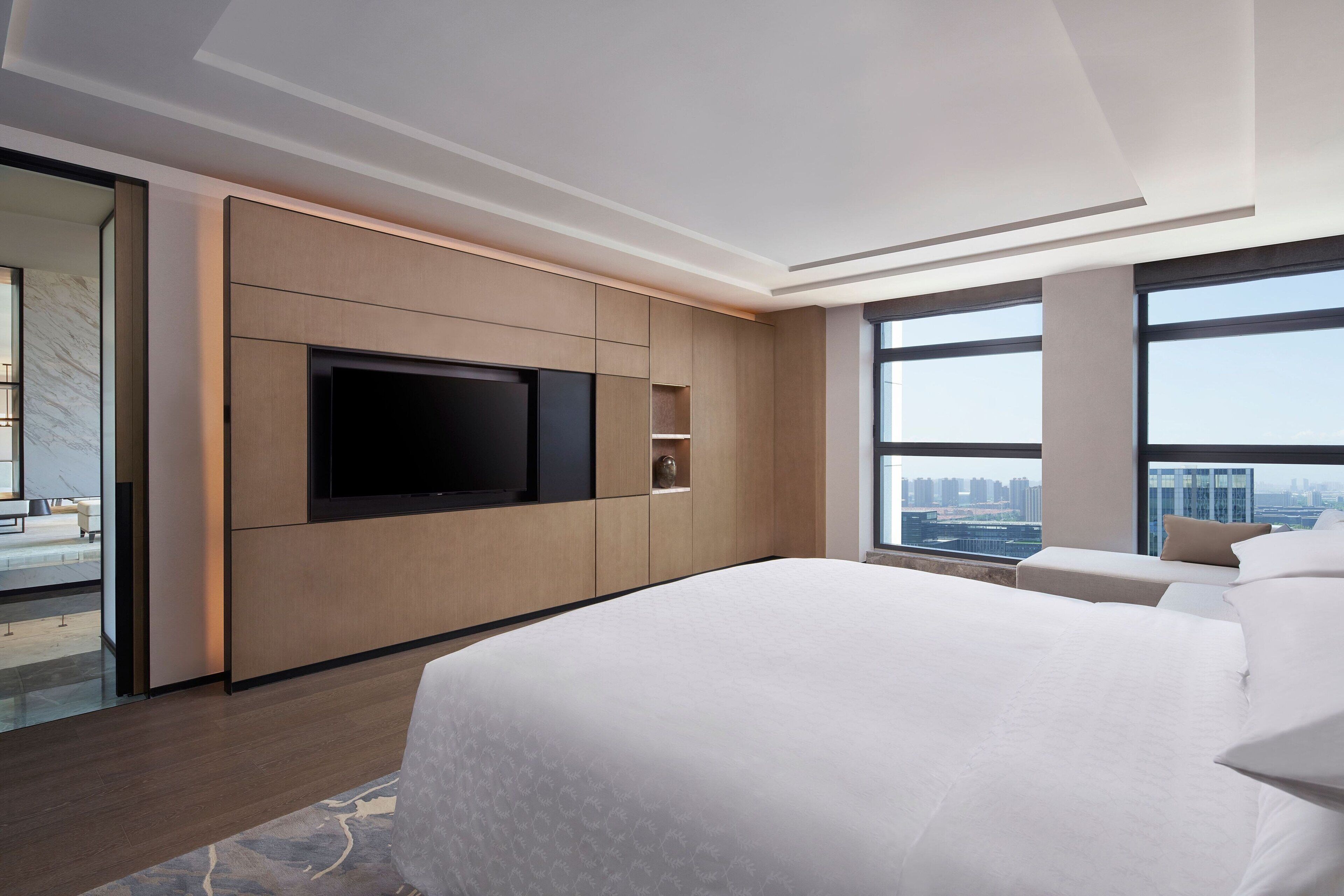 Una habitación de hotel moderna con una cama grande, un televisor de pantalla plana montado en la pared y vistas a la ciudad a través de amplios ventanales.