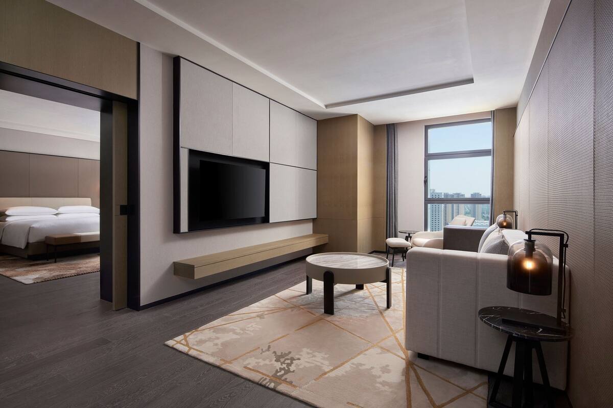 A modern hotel room with a large bed, a flat-screen TV mounted on the wall, a seating area with a sofa and a small table, and a large window offering a city view.