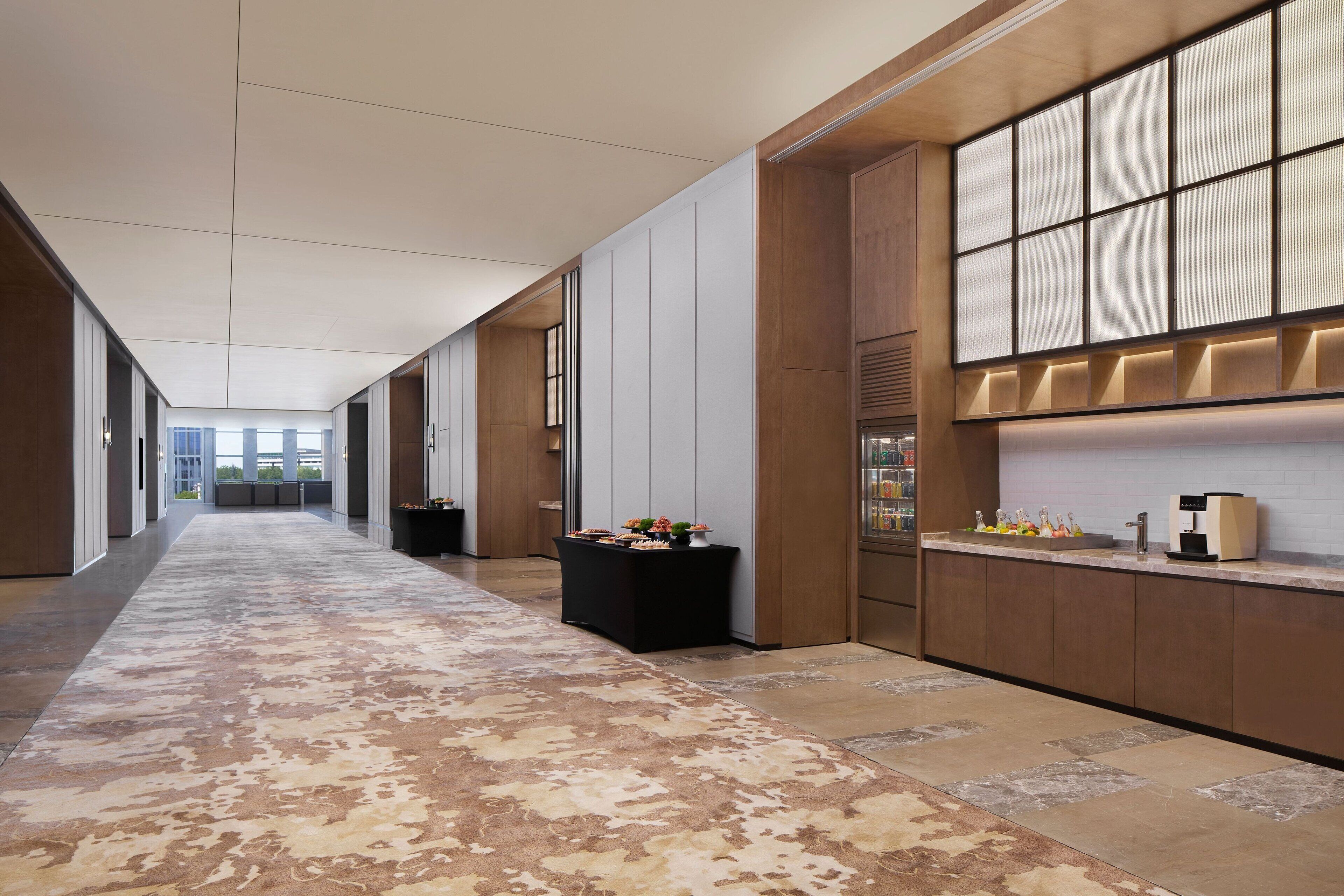 Un pasillo moderno de hotel con alfombra estampada, una recepción con una máquina de café y un ventanal con persianas.
