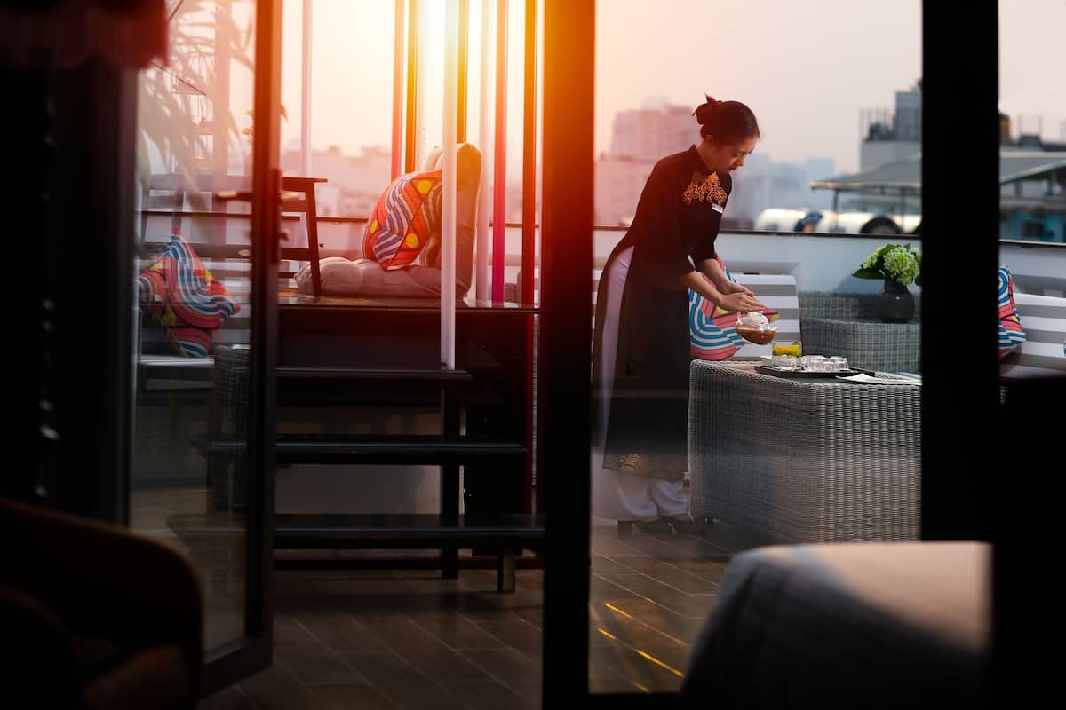 A woman preparing a drink on a balcony with wicker furniture and a city view.