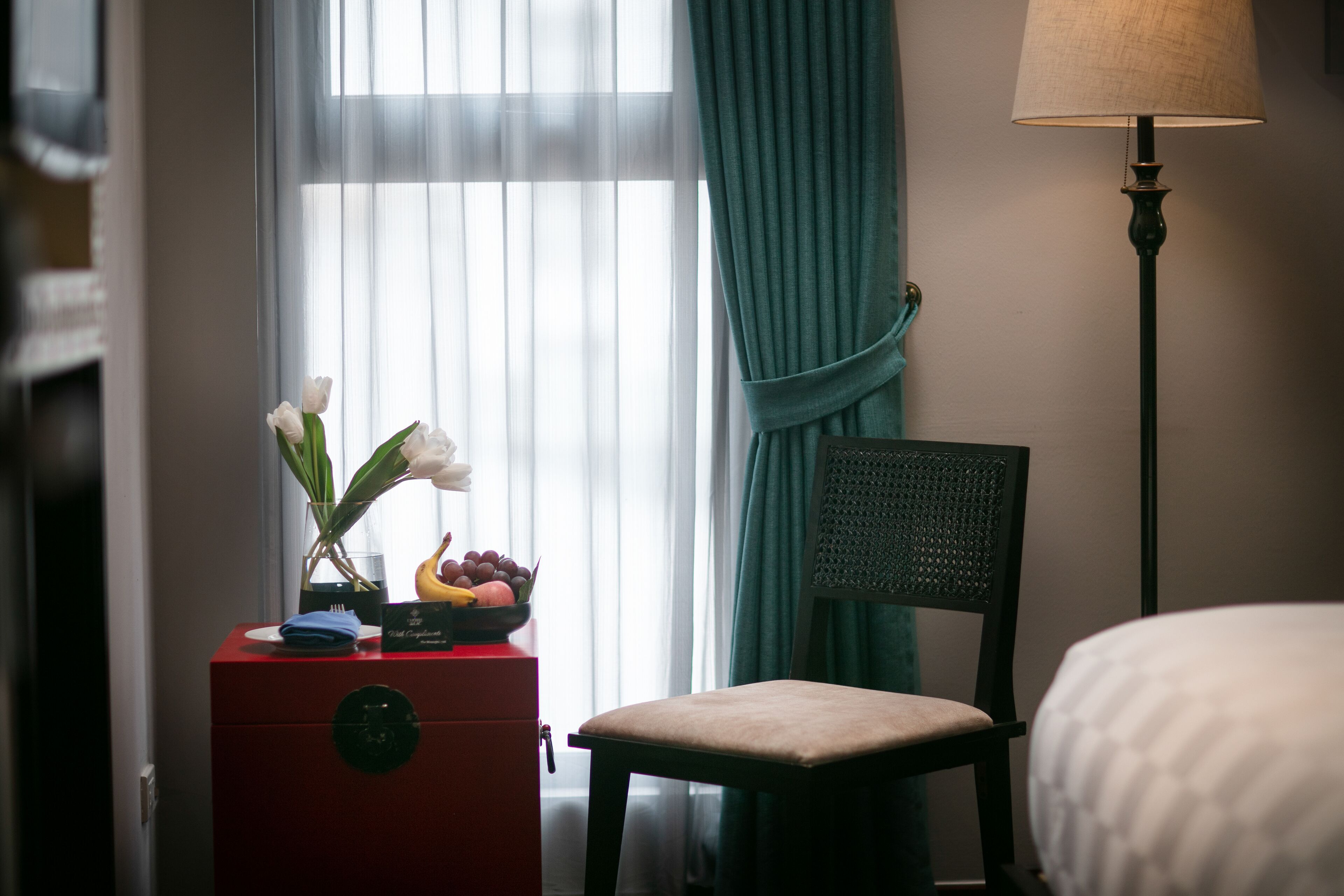 A hotel room with a bed, a chair, a red dresser with a clock, a vase with flowers, and a fruit bowl.