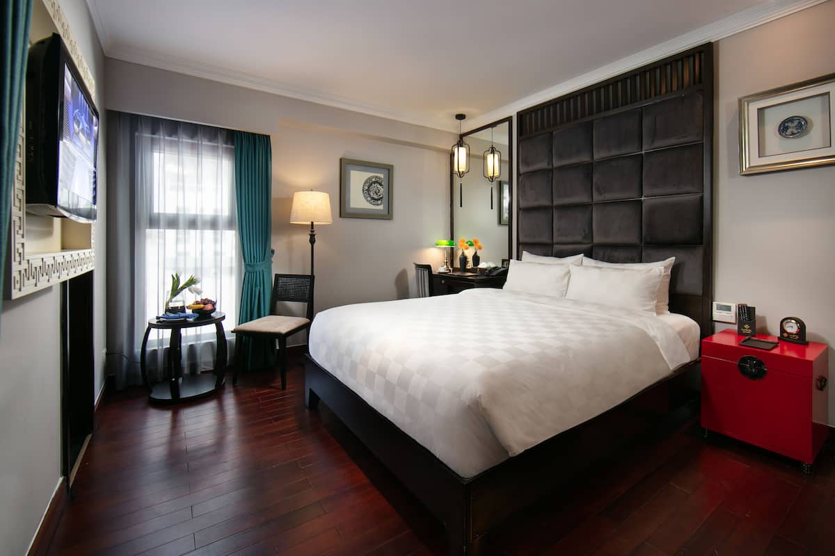 A hotel room with a large bed, a bedside table, a nightstand, a red safe, a wall-mounted TV, a window with curtains, a framed picture, and a floor lamp.