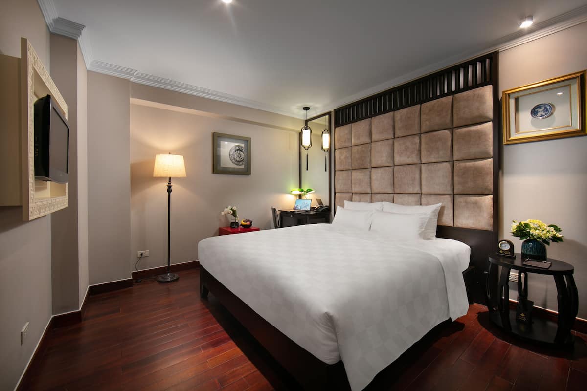 A hotel room with a large bed, a flat-screen TV mounted on the wall, a bedside table with a lamp, and a decorative headboard.