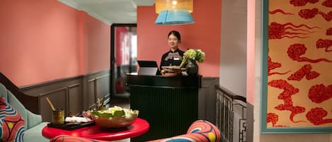 A hotel reception area with a receptionist, a desk with a laptop, a vase with flowers, and a colorful chair.