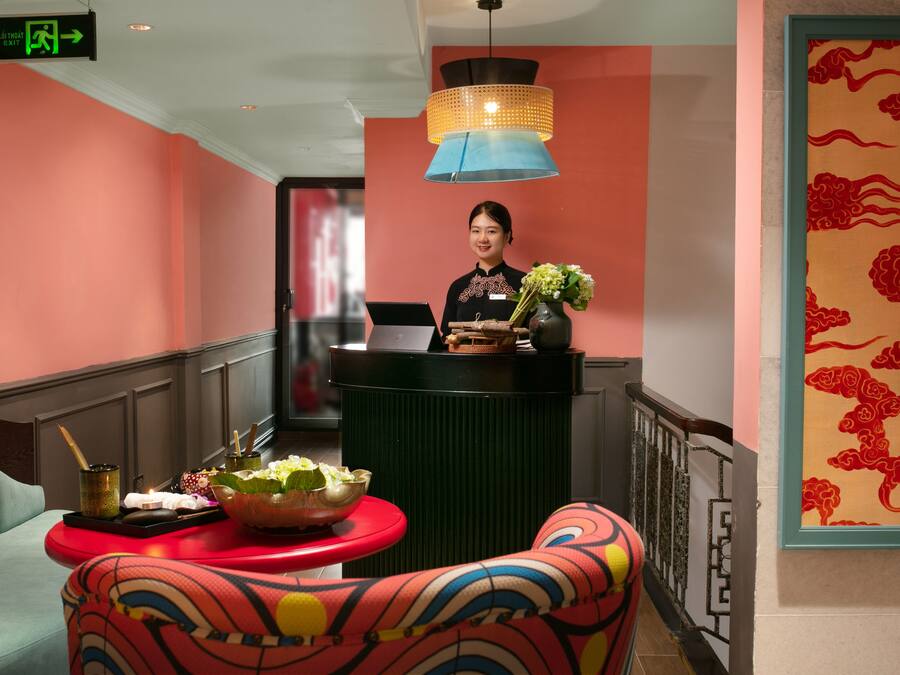 Een hotel receptie met een receptioniste, een bureau met een laptop, een vaas met bloemen en een kleurrijke stoel.