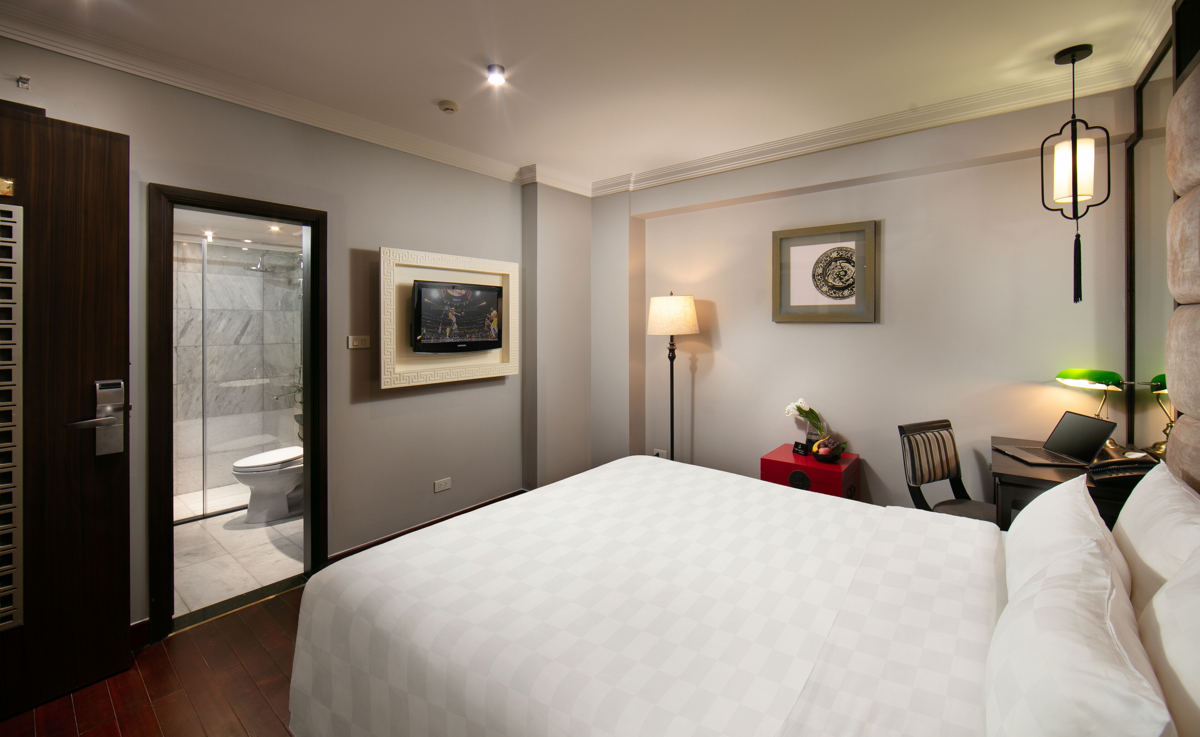 A hotel room with a bed, a TV mounted on the wall, a desk with a laptop, a chair, a bedside table with a lamp, and a framed picture on the wall.