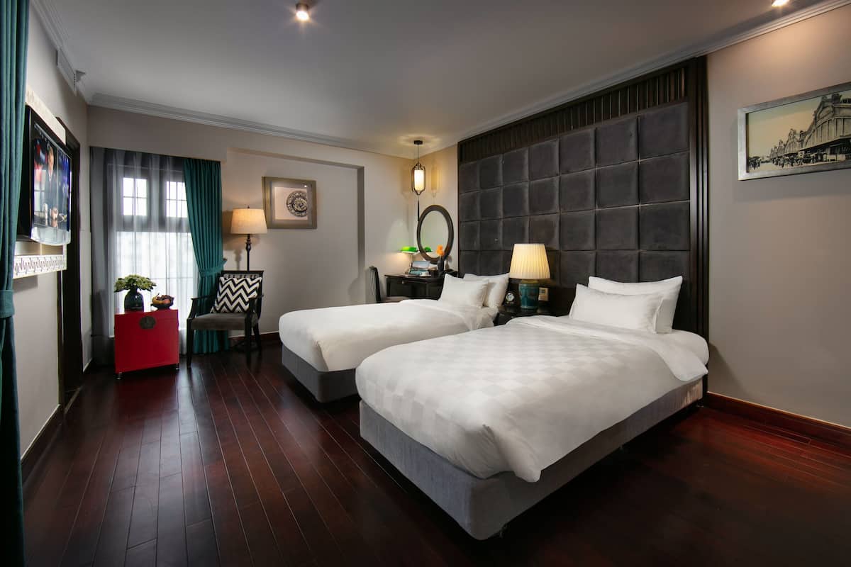 A hotel room with two beds, a red side table, a chair, a mirror, and a framed picture on the wall.