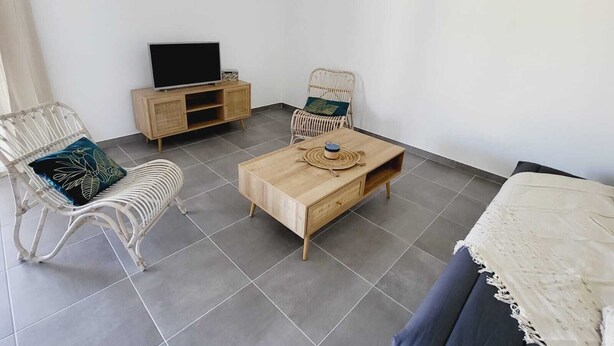 Ein Zimmer mit einem Holztisch, zwei Korbstühlen, einem Fernseher auf einer Holzkommode und einer weißen Wand mit einer Pendelleuchte.
