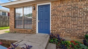 A single-story brick house with a blue front door, a window with blinds, and a small front yard with plants.