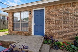 Uma casa térrea de tijolos com porta frontal azul, janela com persianas e um pequeno jardim com plantas.