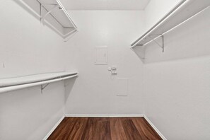 Um quarto pequeno com paredes brancas, soalho de madeira, duas prateleiras brancas e uma porta branca.
