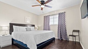 A bedroom with a bed, a nightstand, a chair, a ceiling fan, and a window with curtains. - World Cup Walkable | King Bed Retreat (Arlington)