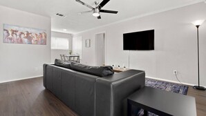 A modern living room with a gray sofa, a ceiling fan, a flat-screen TV, and a dining area in the background.