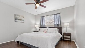 A bedroom with a bed, a ceiling fan, a window with curtains, and a bedside table with a lamp.