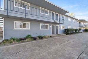 Um prédio de apartamentos com dois andares, varanda, entrada de concreto e um pequeno jardim.
