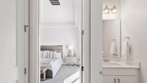 Habitación de hotel moderna con una cama bien hecha, una mesita de noche con lámpara, un espejo grande sobre el lavabo y una decoración en blanco y colores limpios.