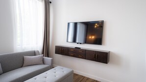 A modern living room with a grey sofa, a flat-screen TV mounted on a dark wooden console, and sheer curtains on the window. - Mount Alverno Luxury Resorts (Caledon)