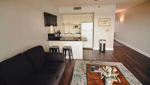 A living room with a dark-colored sofa, a coffee table with a vase of flowers, and a kitchen area with white cabinets and a microwave.