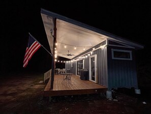 A small, single-story house with a wooden deck, an American flag, and string lights. - Shipping Container Cabin Couples Paradise! (Rocky Point)