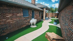 A brick building with a curved pathway leading to a door, surrounded by green artificial grass and a large rock. - Spark Getaways: The Mulligan Spa and Golf (Lubbock)