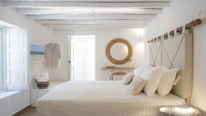 A bedroom with a large bed, a bedside table, a wall-mounted mirror, and a window with a view of the beach. - Pietra Bianca 1891 Suites Mykonos (Mykonos)