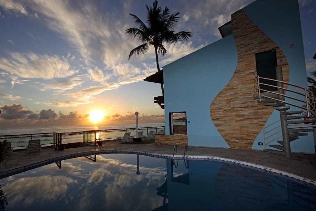 Un espace aménagé au bord de la piscine, offrant une vue sur le coucher de soleil sur l’océan, un palmier et un bâtiment moderne doté d’une façade en bois.