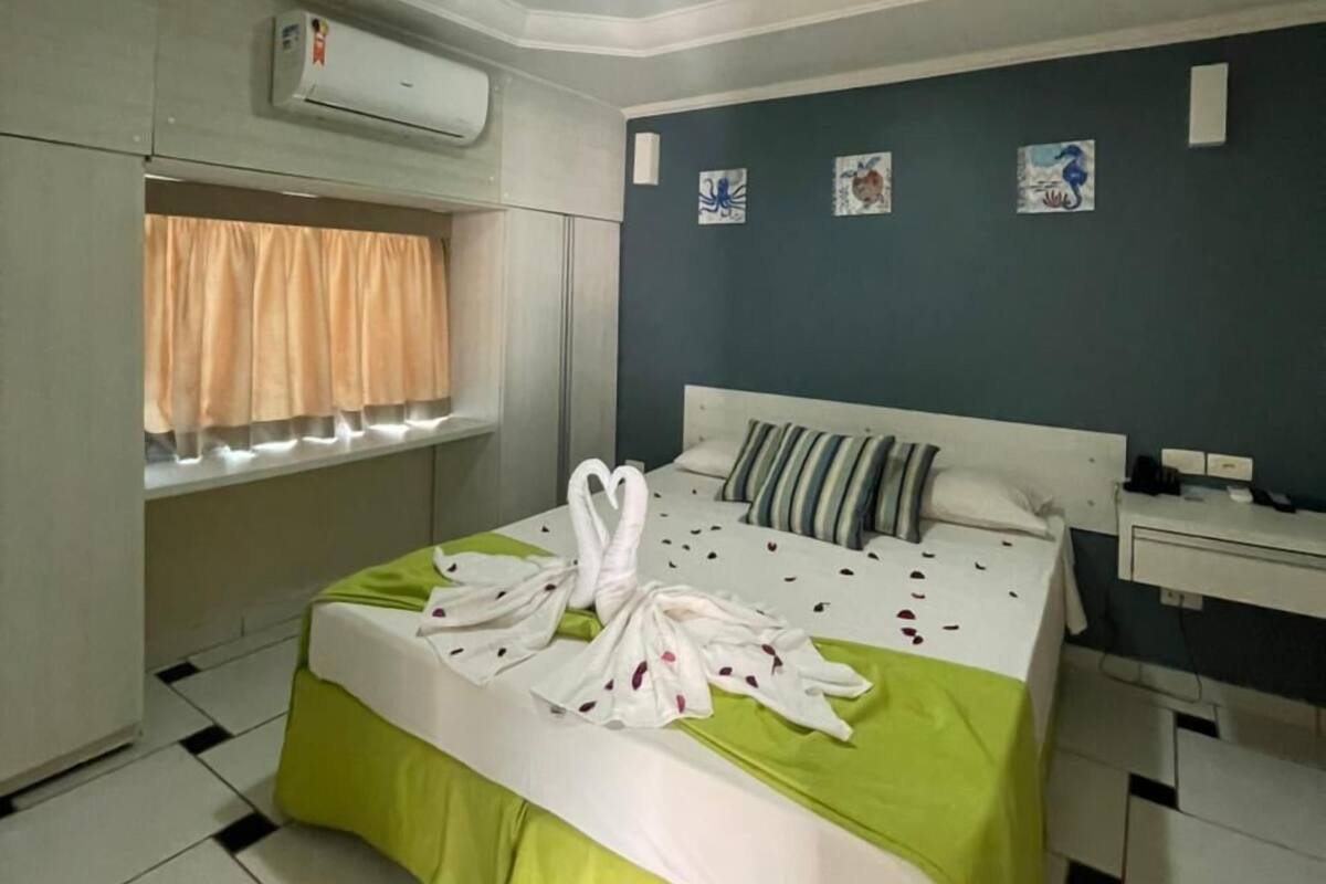 A hotel room with a bed, a window with curtains, an air conditioning unit, and a bedside table.