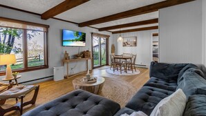 A spacious living room with a large sofa, a dining area with a wooden table and chairs, a television mounted on the wall, and large windows offering a view of the outdoors.