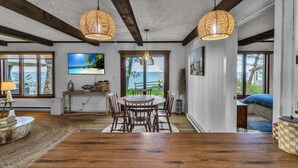 A dining area with a wooden table and chairs, two hanging wicker lamps, a television on the wall, and a view of the outdoors through large windows.