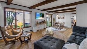 A spacious living room with a large window, a flat-screen TV, and a dining area with a table and chairs.