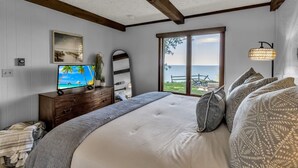 A bedroom with a large bed, a TV, a wooden dresser, a window with a view of the sea, and a lamp.