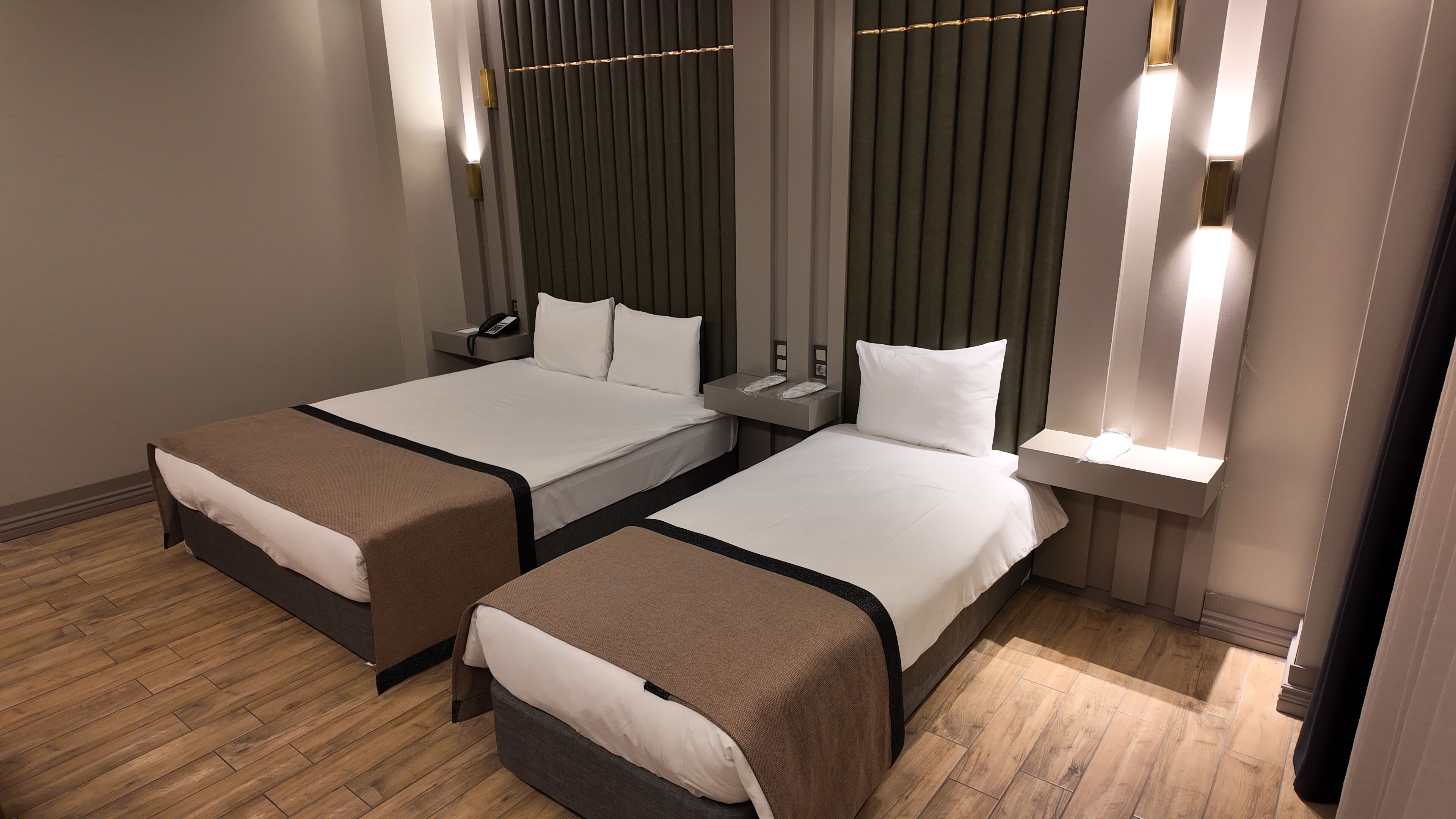 Habitación de hotel con dos camas, piso de madera, mesita de noche con teléfono e iluminación en la pared.