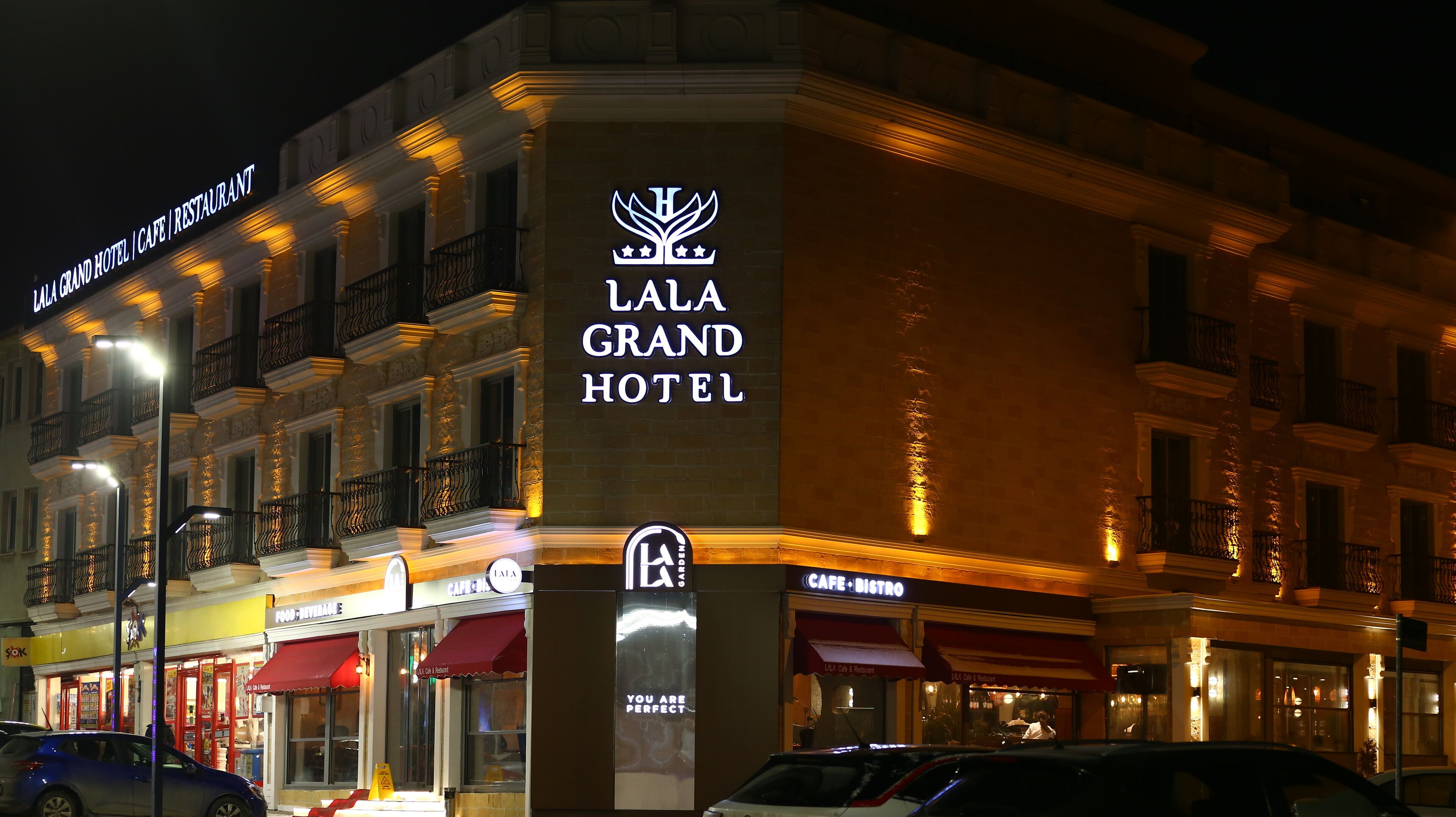 Het gevel van het Lala Grand Hotel 's nachts, met verlichte signage en aangrenzende cafés.
