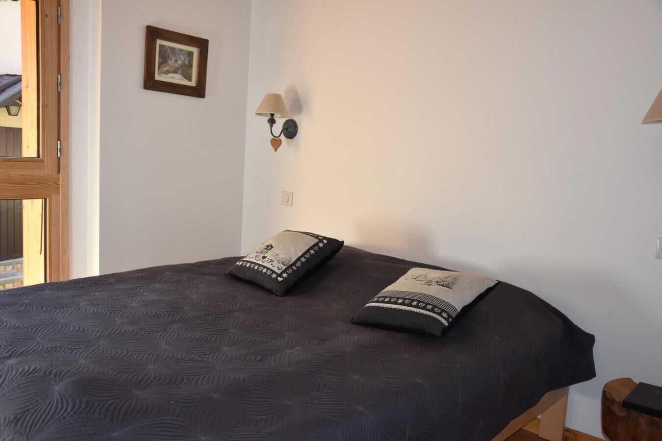 Un lit recouvert d  ’une couvre-lit à motifs sombres, deux oreillers ornés de motifs décoratifs, un luminaire fixé au mur et un tableau encadré accroché au mur.