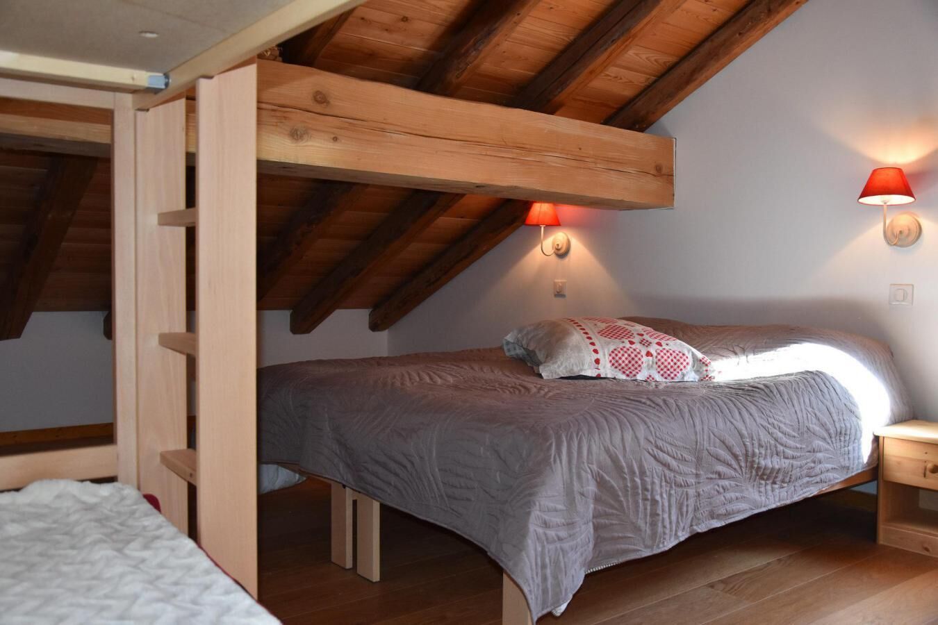 Une chambre à coucher aménagée dans un espace sous les toits, avec un plafond en pente, des poutres en bois, un lit recouvert d’une couverture grise, une lampe rouge et une petite table de chevet en bois.