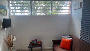 A living room with a brown sofa, a small table with a picture frame and a calendar, a vase with dried plants, a TV mounted on the wall, and a window with blinds.