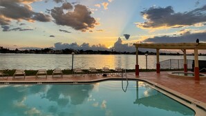Une piscine dotée de chaises longues et d’une pergola en bois, offrant une vue sur une étendue d’eau avec un coucher de soleil en arrière-plan.
