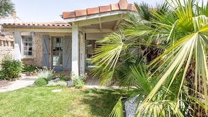 Uma casa de pedra com telhado em telhas, varanda coberta e um jardim bem cuidado com palmeiras e flores.