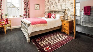A bedroom with a four-poster bed, a nightstand, a bedside table, a window with curtains, a floral wallpaper, and a red towel on a rack. - 1 bedroom accommodation in Kelso (Kelso)