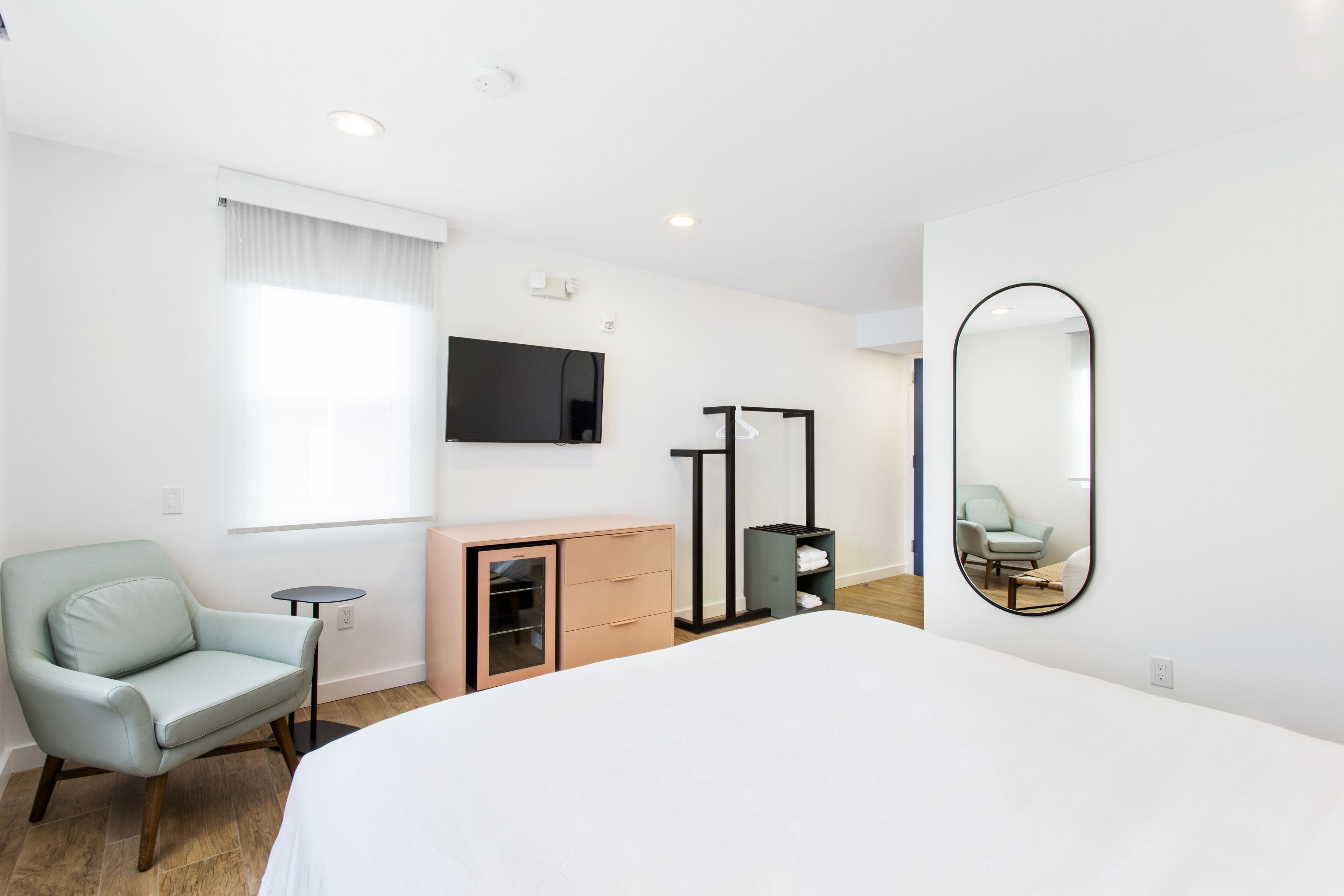 Ein modernes Hotelzimmer mit einem großen Bett, einem hellblauen Sessel, einem kleinen Tisch, einem an der Wand befestigten Fernseher und einem Spiegel.