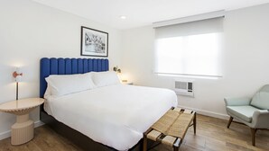 Ein modernes Schlafzimmer mit einem großen Bett, einem blauen Kopfteil, einem Nachttisch, einem Sessel und einem Fenster mit Jalousien.