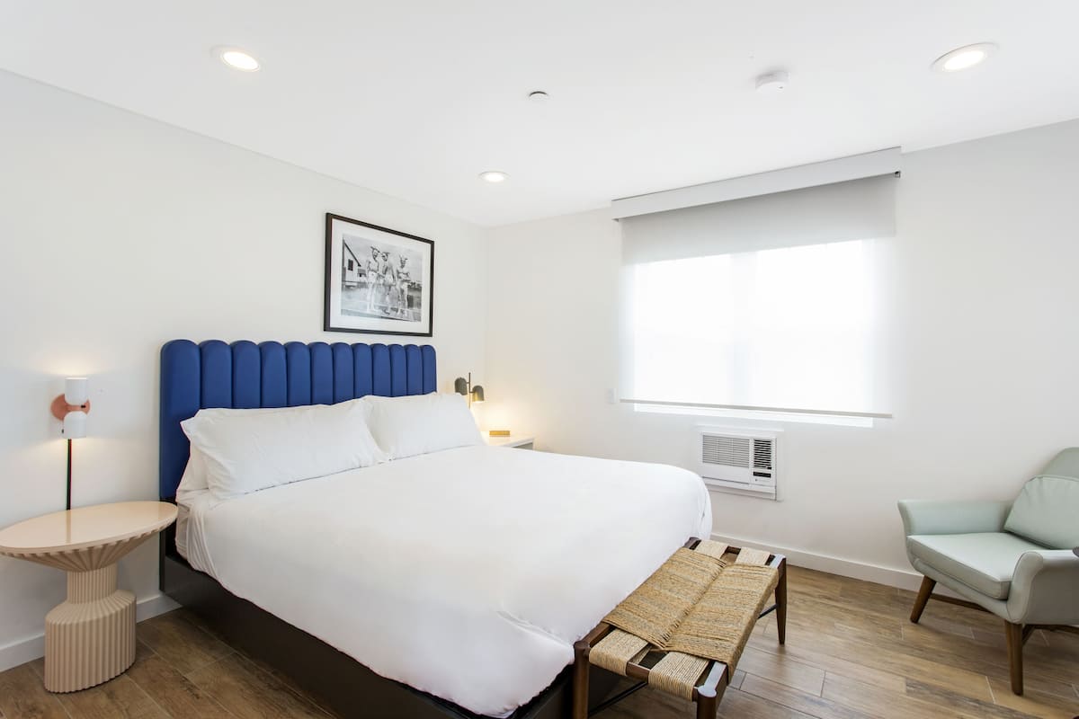 A modern bedroom with a large bed, a blue headboard, a bedside table, a chair, and a window with blinds.