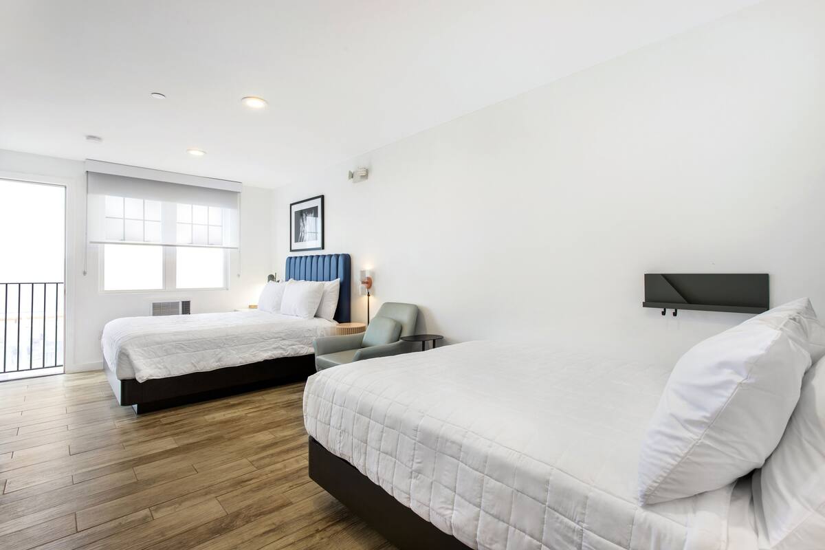 A hotel room with two beds, a chair, a desk, and a large window.