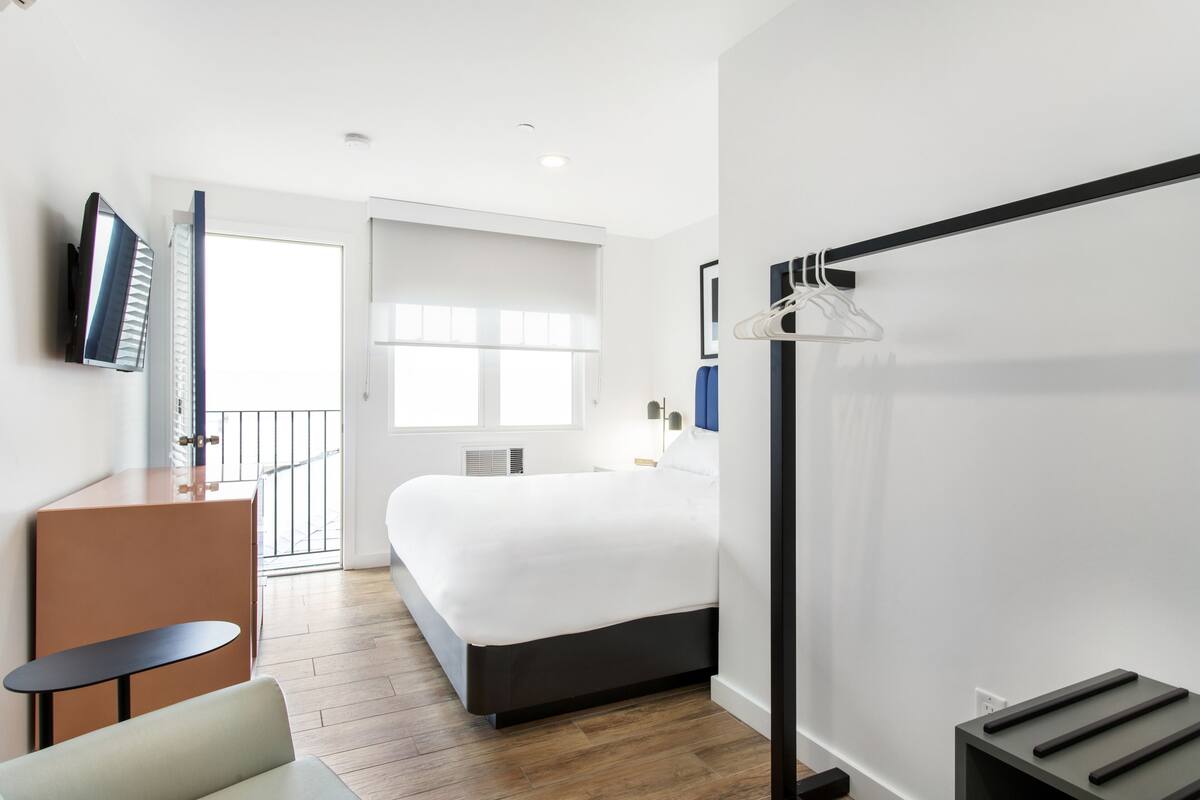 A hotel room with a bed, a desk, a chair, a TV, and a coat rack.