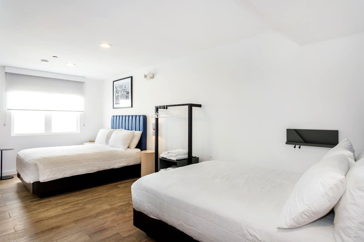 A hotel room with two beds, a bedside table, a chair, a TV mounted on the wall, and a window with blinds.
