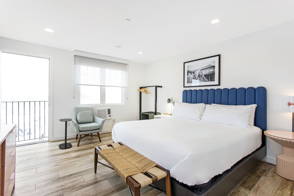 A modern hotel room with a large bed, a seating area with a bench, a small table, and a chair.